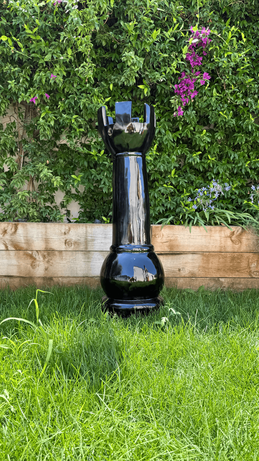 Statue Échecs - GRANDIOSE en forme de tour noire, 1m40 en résine, posée sur pelouse devant haie fleurie. Design contemporain pour jardin décoratif.