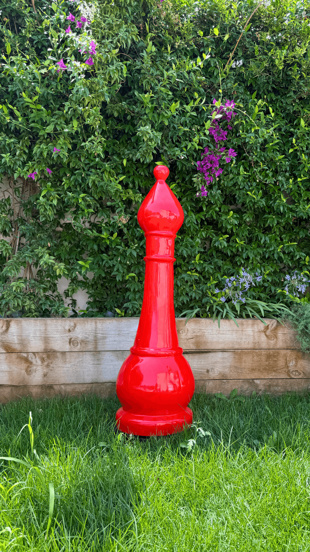 Statue Échecs - GRANDIOSE en résine rouge représentant un fou géant de 1m40, posée sur pelouse devant un mur végétal. Idéale pour jardin décoratif.
