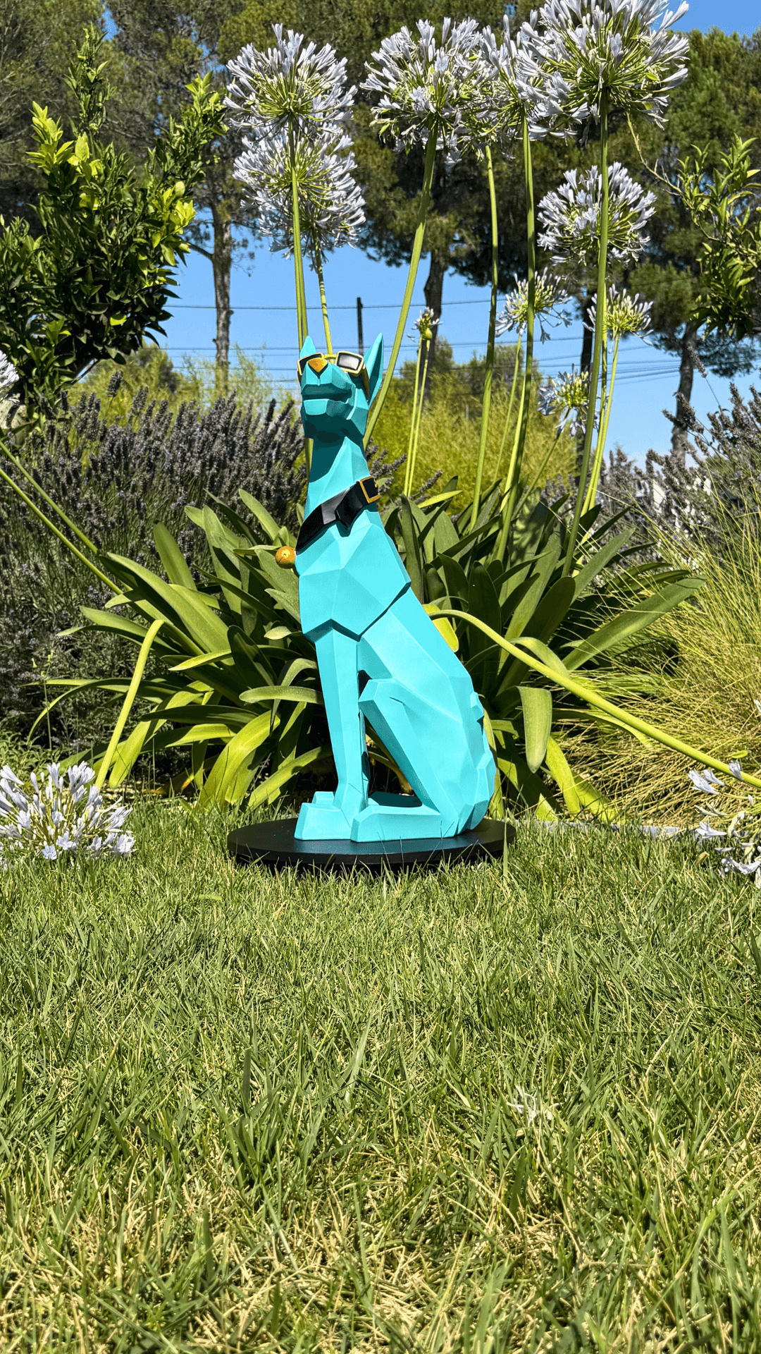 Statue Chien Majordome - Design Moderne Déco Luxe en résine turquoise posée dans un jardin fleuri, parfaite pour une déco extérieure originale.
