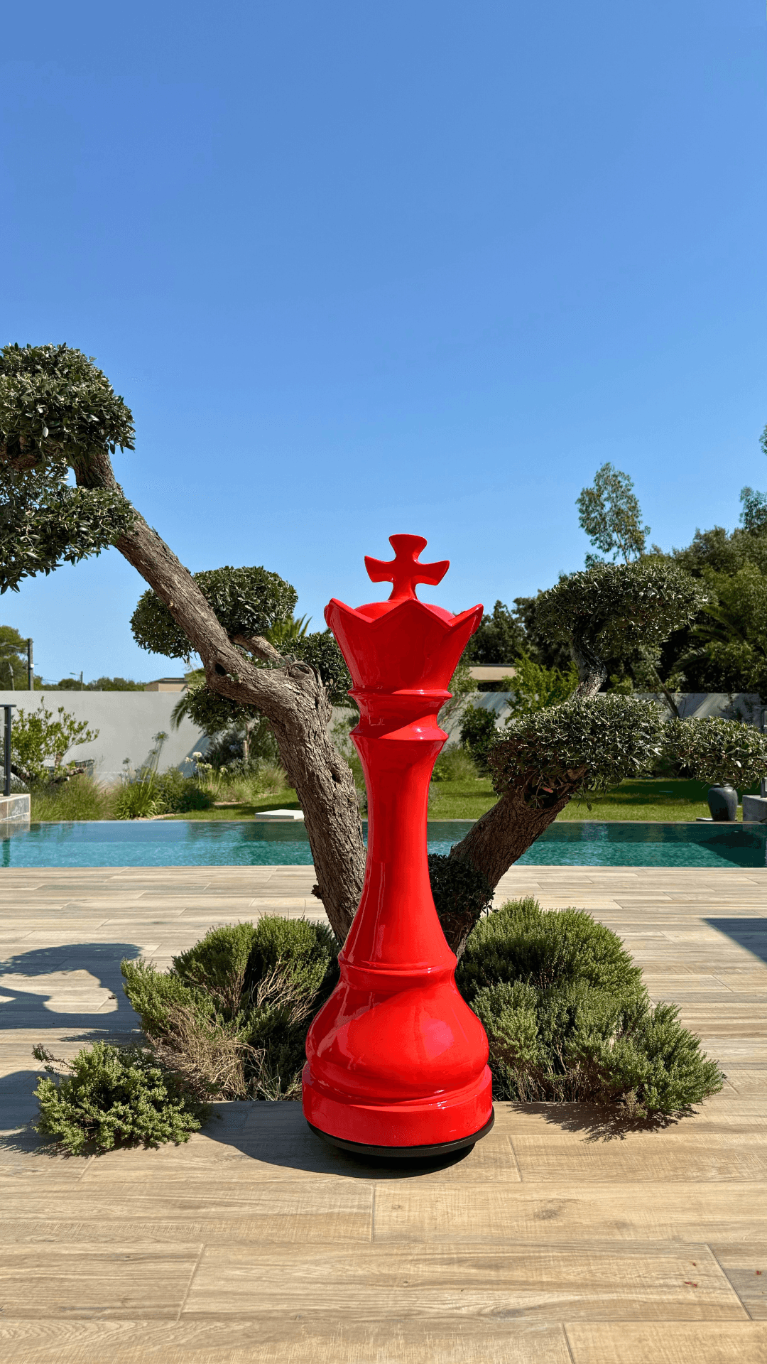 Statue d’échecs géante rouge en résine représentant une reine, modèle GRANDIOSE, posée sur une terrasse extérieure près d’un arbre taillé. Idéale pour décoration de jardin ou espace public.
